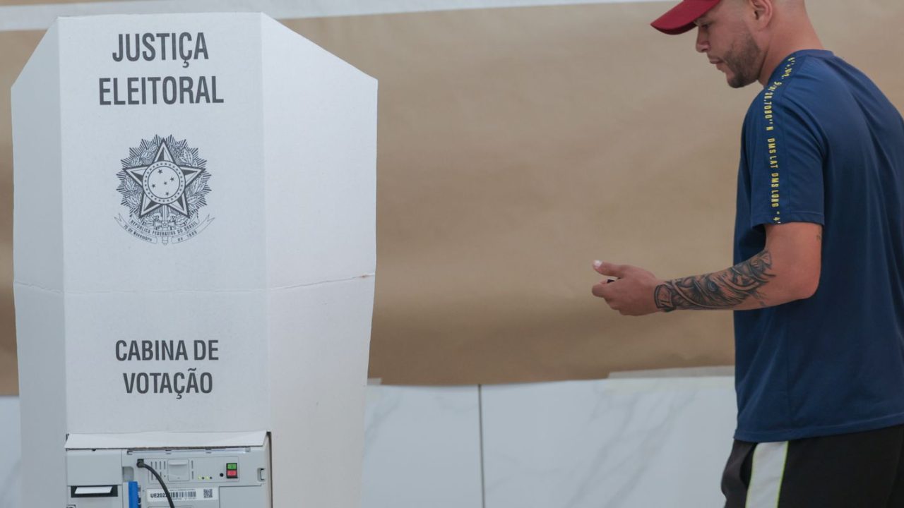 Eleitor segurando papel com números de candidatos ao lado da urna eletrônica